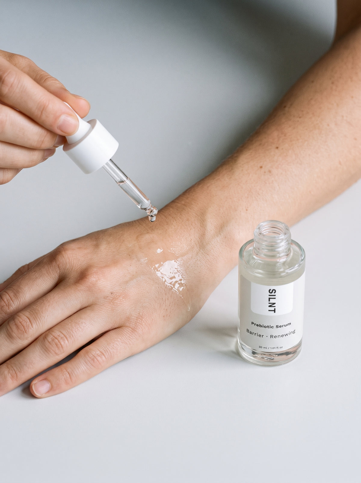 Hand applying SILNT Prebiotic Serum from the dropper onto the forearm, bottle beside it