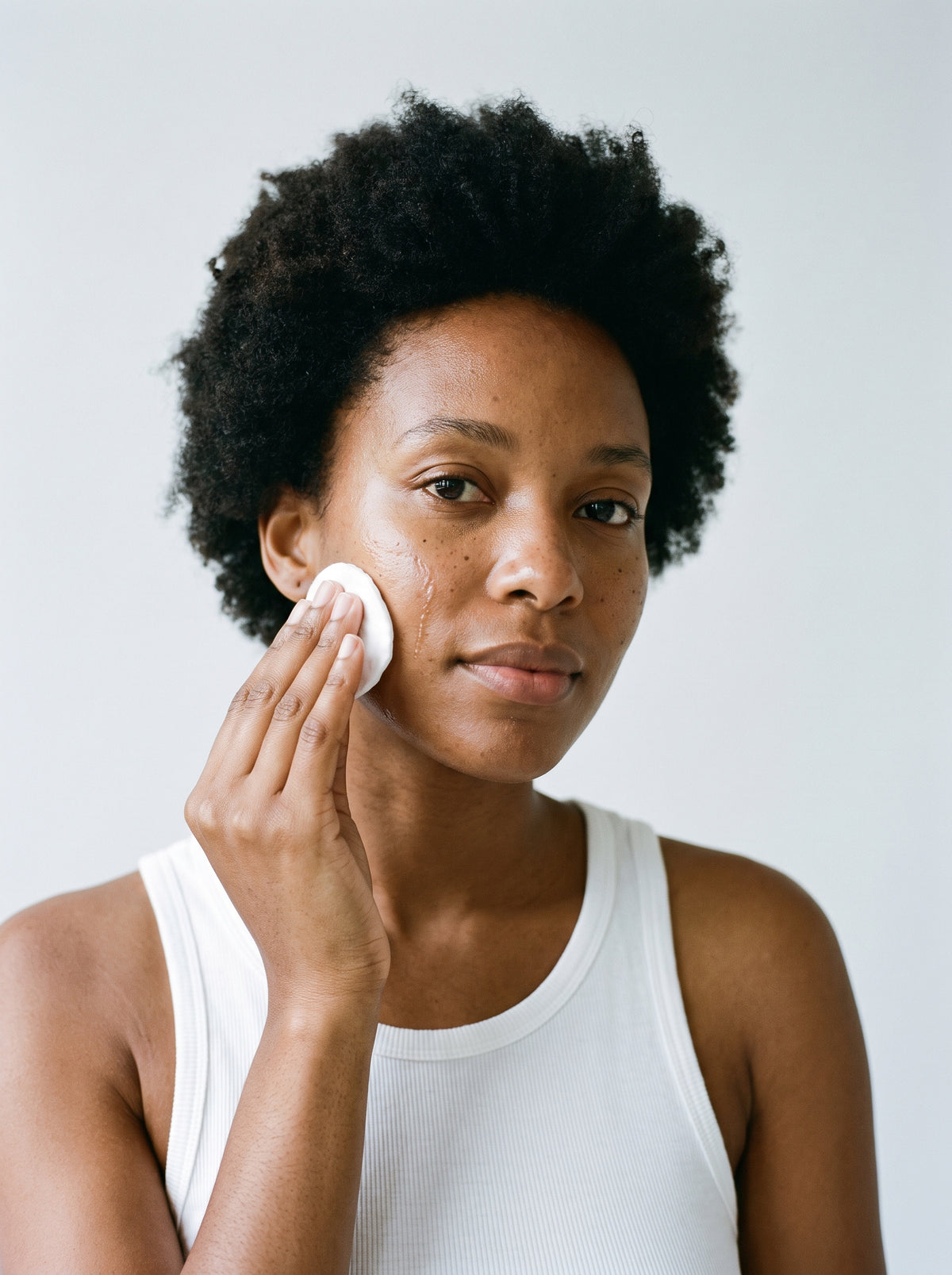 Woman in a white tank top sweeping SILNT Glycolic Toner over her cheek with a cotton pad