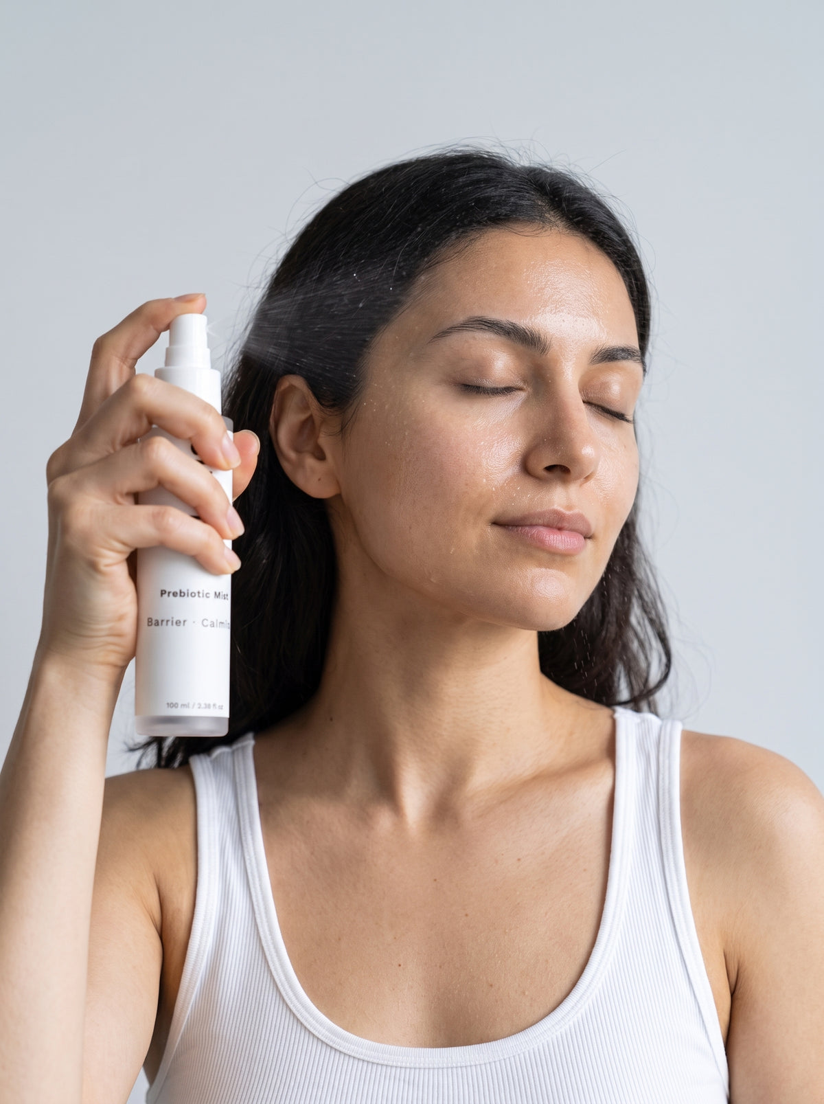 Woman in a white tank top spraying SILNT Prebiotic Mist onto her face with eyes closed