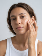 SILNT Facial Oil pressed into a model's cheek with her fingertips, white tank top, light grey background