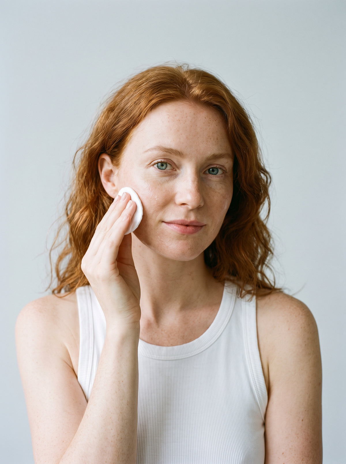 Red-haired woman in a white tank top applying SILNT Glycolic Toner with a cotton pad