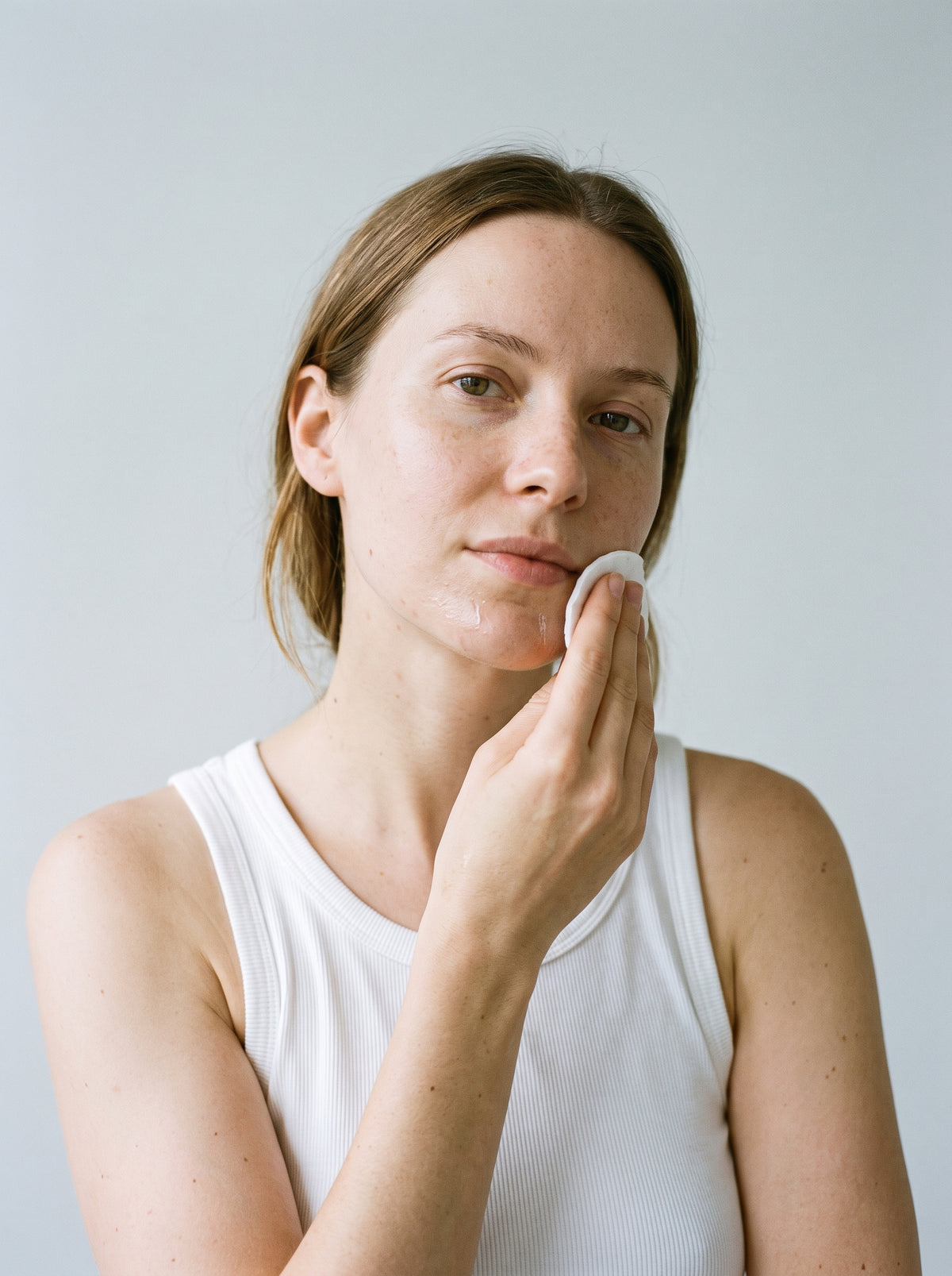 SILNT Makeup Remover swept over a model's cheek with a cotton pad, white tank top, light grey background
