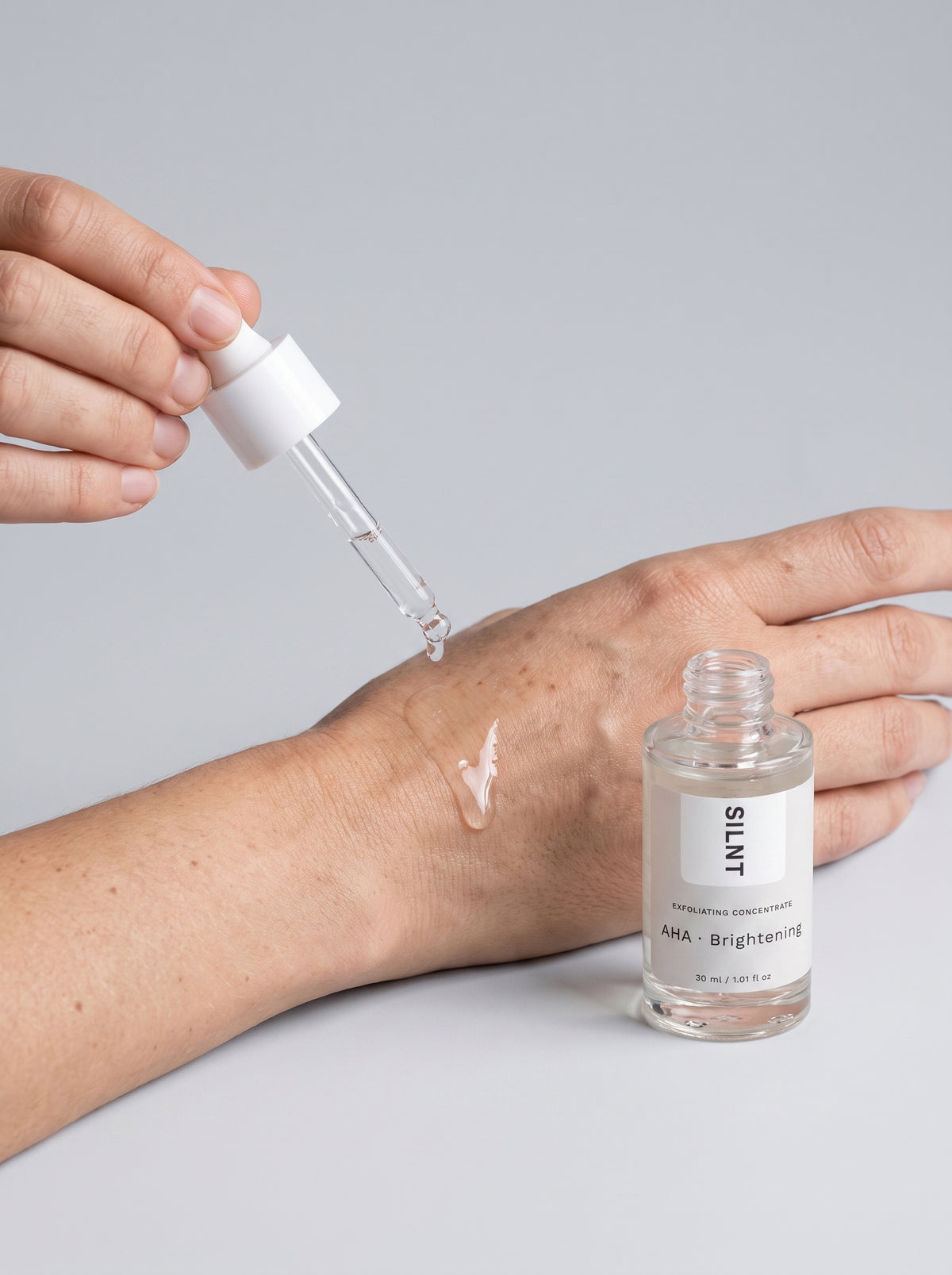 SILNT Exfoliating Concentrate clear gel dropped onto the back of the hand from the glass pipette, light grey background