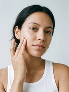 SILNT Cleansing Oil massaged over a model's cheek with her fingers, white tank top, light grey background