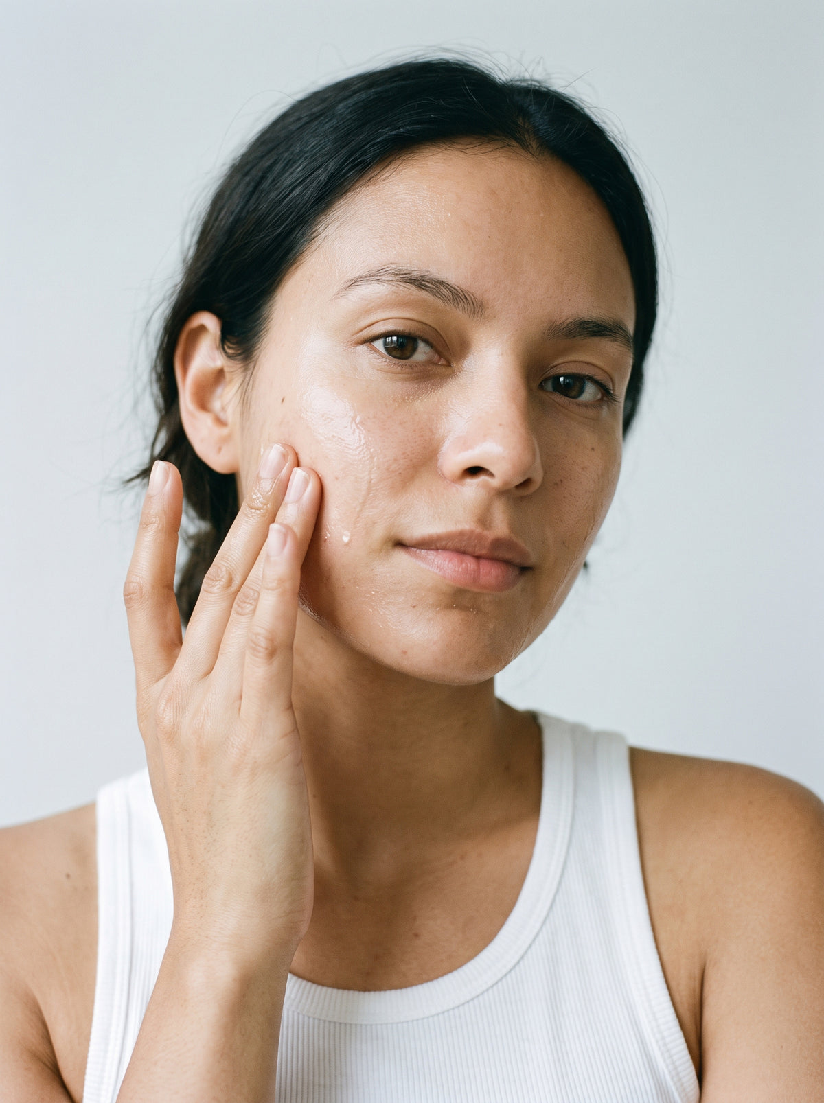 SILNT Cleansing Oil massaged over a model's cheek with her fingers, white tank top, light grey background