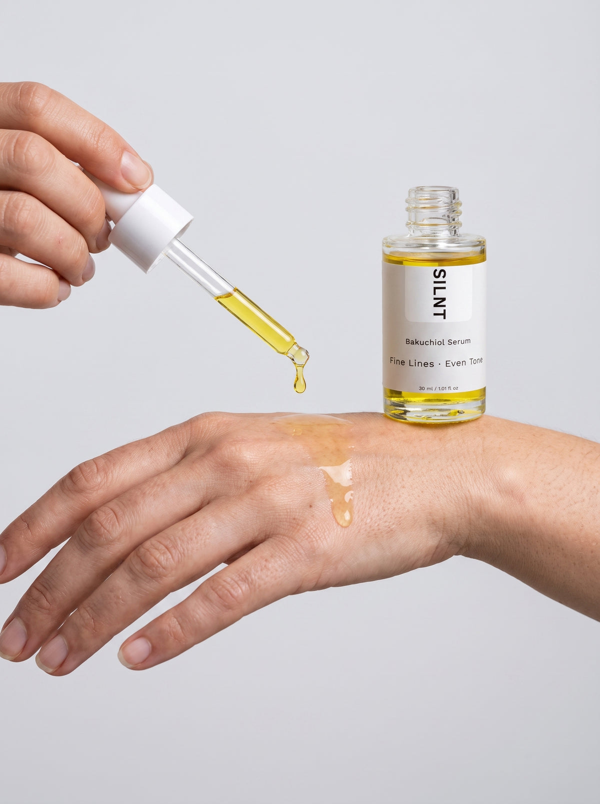 SILNT Bakuchiol Serum golden oil dropped onto fingertips from the glass pipette, light grey background