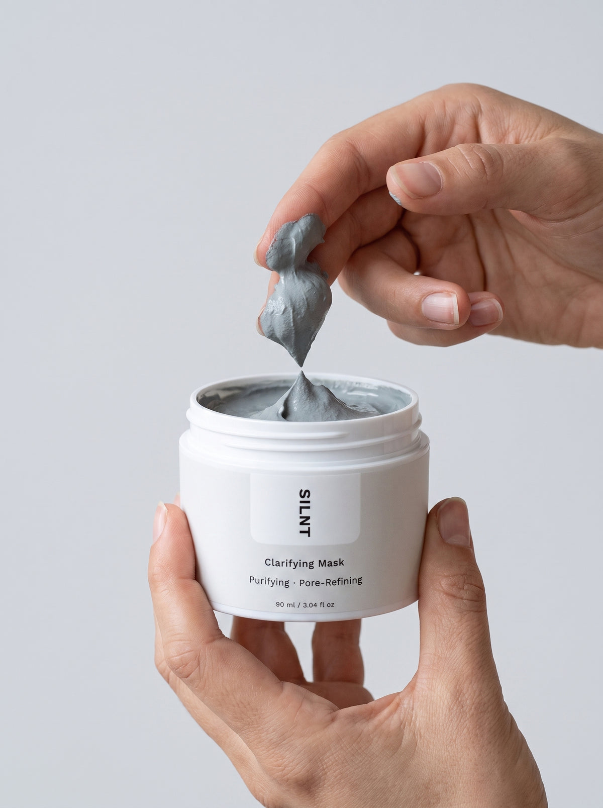 SILNT Clarifying Mask grey clay lifted from the open jar with fingertips, light grey background