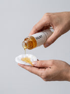 SILNT Makeup Remover poured from the bottle onto a cotton pad held in one hand, light grey background