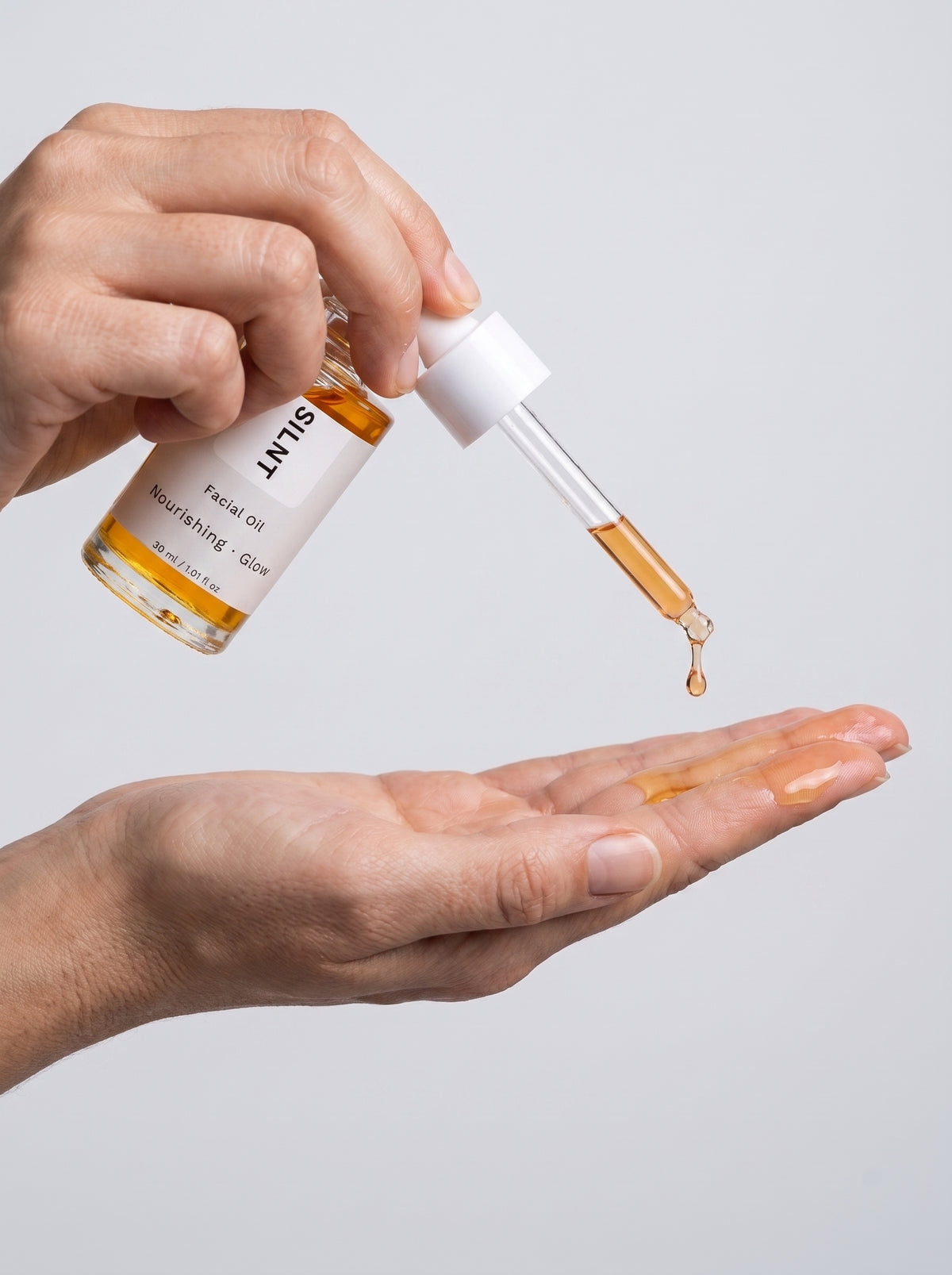 SILNT Facial Oil amber oil dropped onto fingertips from the glass pipette, light grey background