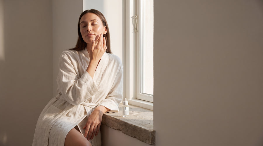 A woman in a linen robe sitting by a window in morning light, eyes closed, with SILNT Prebiotic Serum on the windowsill