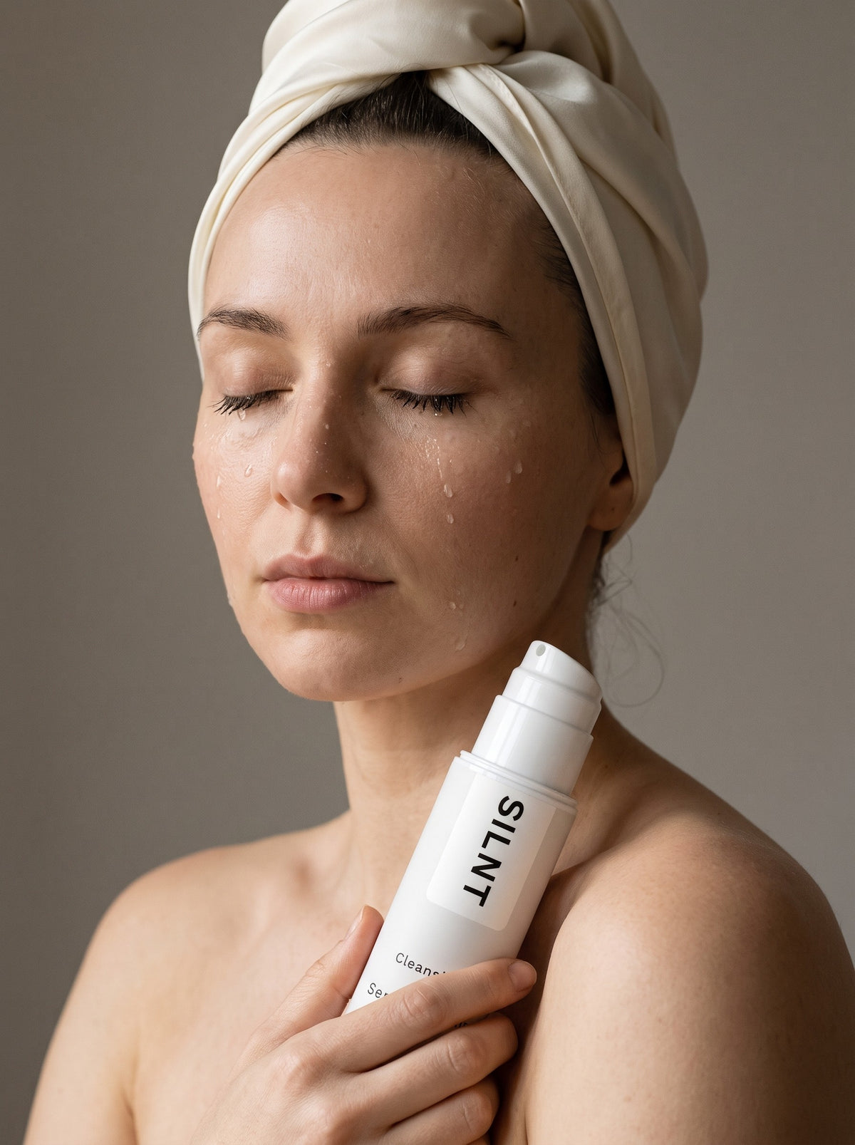 A woman with her eyes closed and water on her skin, a silk scarf in her hair, holding SILNT Cleansing Oil